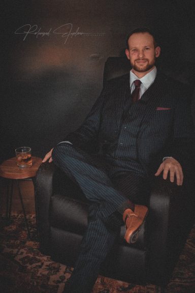 A well-dressed man in a suit sits comfortably in a chair, with a drink on a nearby table.