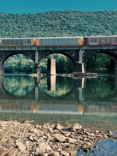 Train crossing a bridge over a calm river reflecting the surrounding hills.