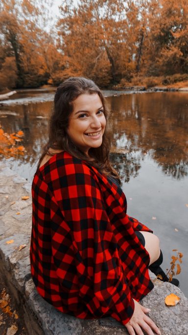 Woman in a red and black checkered sweater sits by a river surrounded by autumn foliage.
