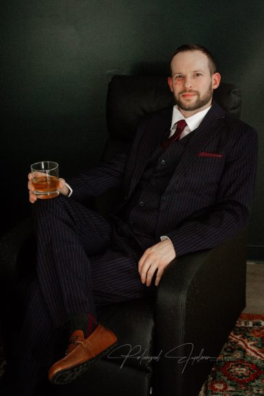 Man in a dark suit holding a drink, seated in a black armchair against a dark background.